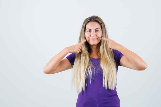 紫のTシャツで彼女の笑顔を指して、陽気な正面図を探しているブロンドの女性。