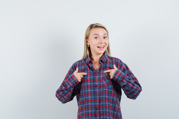 Blonde woman pointing at herself with index fingers in checked shirt and looking excited. front view.