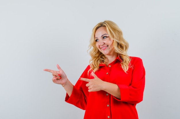 Blonde woman pointing left with index fingers in red blouse and looking happy.