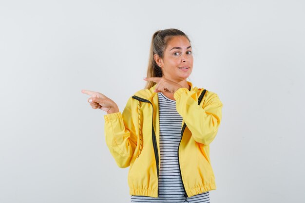 Blonde woman pointing left with index fingers in yellow bomber jacket and striped shirt and looking pretty