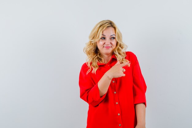Blonde woman pointing right side with index finger in red blouse and looking happy.
