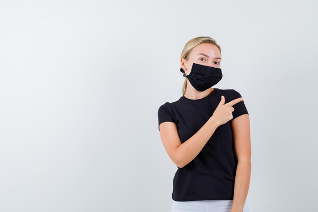 Blonde woman pointing right with index finger in black t-shirt, white pants