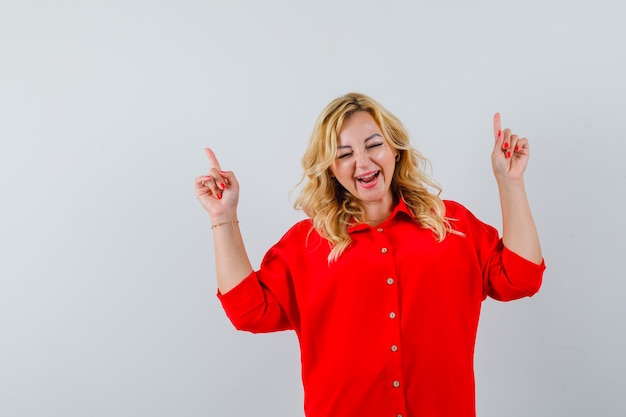 Free photo blonde woman pointing up with index fingers in red blouse and looking happy.