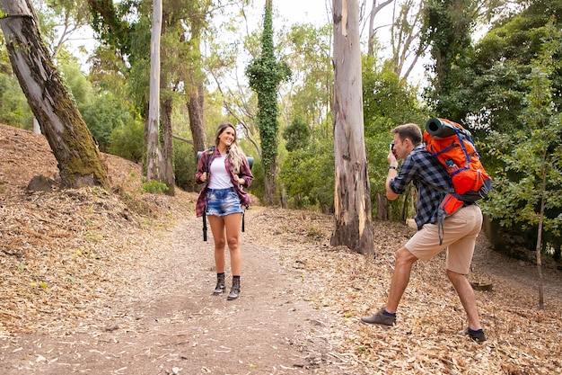 Blonde woman posing for photo on road in forest. Caucasian man holding camera and shooting on nature. Two happy people trekking with backpacks. Tourism, adventure and summer vacation concept