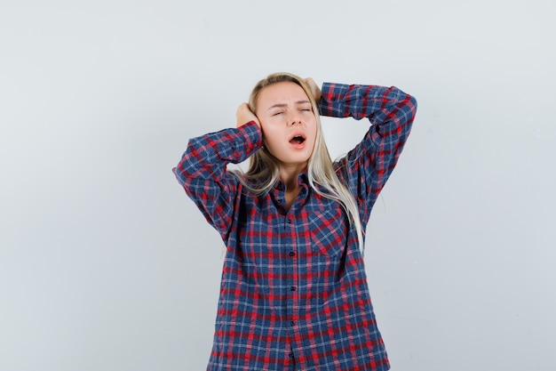 Blonde woman pressing hands on ears, standing with open mouth in checked shirt and looking exhausted , front view.