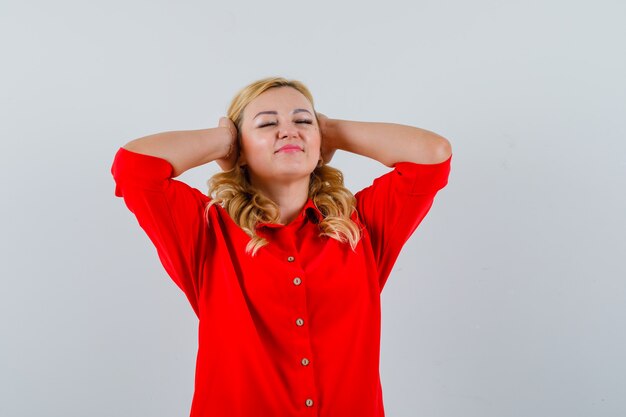 Blonde woman pressing hands on ears while closing eyes in red blouse and looking happy.