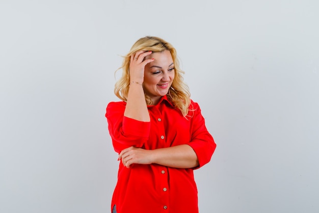 Blonde woman putting hand on head, looking away and posing at camera in red blouse and looking happy.