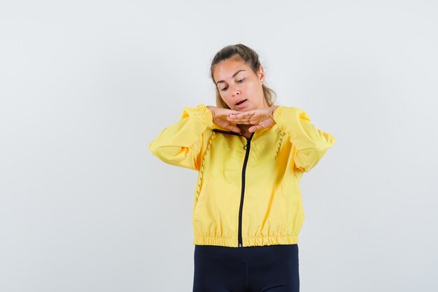 Blonde woman putting hands under chin in yellow bomber jacket and black pants and looking focused