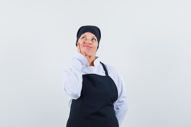 Foto gratuita donna bionda che mette il dito indice sulla guancia, pensando a qualcosa in uniforme nera del cuoco e che sembra carina