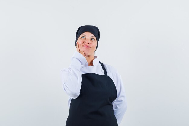 Blonde woman putting index finger on cheek,thinking about something in black cook uniform and looking pretty