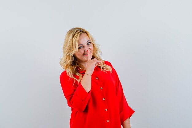 Blonde woman putting index finger on mouth, showing silence gesture in red blouse and looking happy