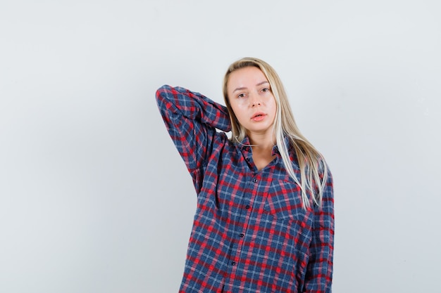 Foto gratuita donna bionda che mette un braccio dietro la testa, che propone alla macchina fotografica in camicia a quadri e che sembra attraente. vista frontale.