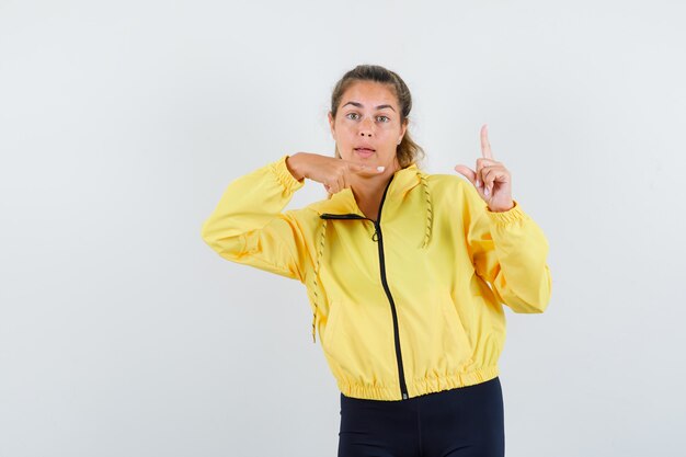 Blonde woman raising index finger in eureka gesture, pointing to it in yellow bomber jacket and black pants and looking happy