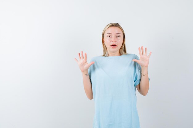 Blonde woman raising palms in surrender gesture in blue t-shirt and looking surprised