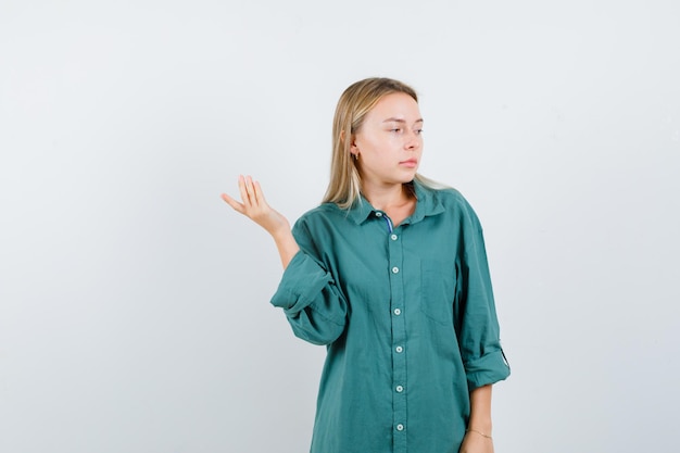 Blonde woman raising spread palm while looking aside in green shirt and looking pensive.