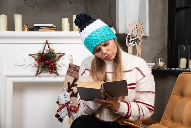 Blonde woman reading interesting book near the fireplace. 