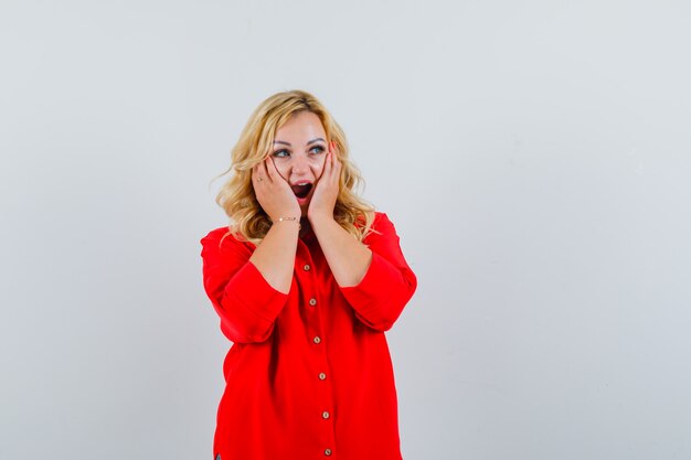 Blonde woman in red blouse holding hands on cheek and posing at camera and looking surprised