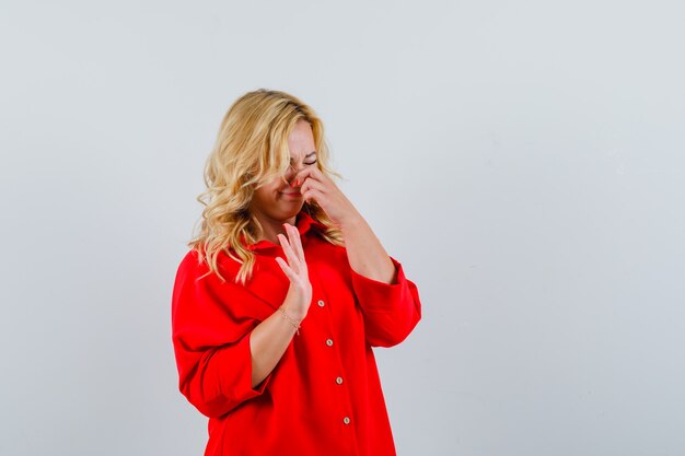 Blonde woman in red blouse pinching nose due to smell and looking annoyed,