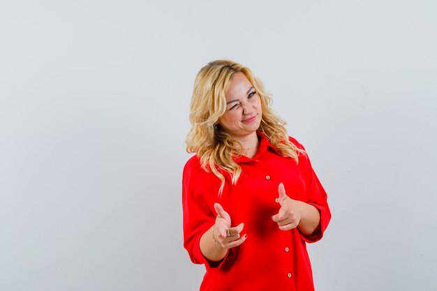 Blonde woman in red blouse pointing index fingers at camera, winking and looking happy