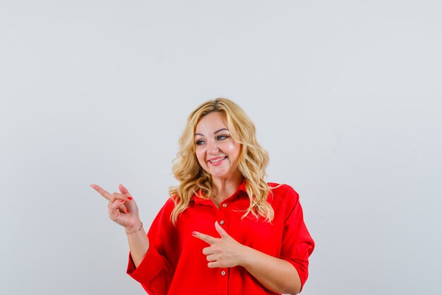 Blonde woman in red blouse pointing left with index fingers and looking happy
