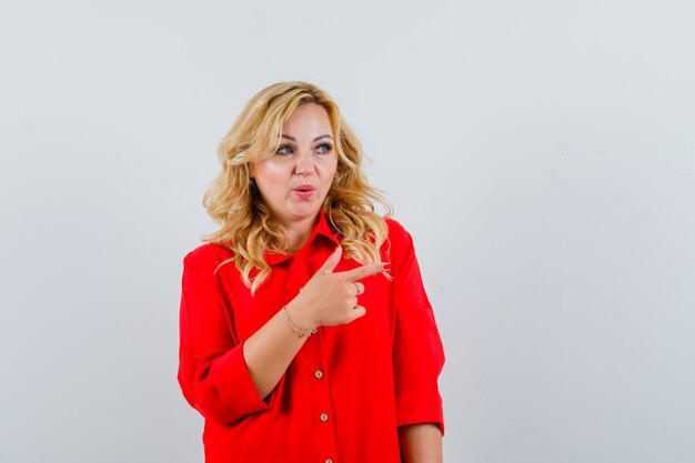 Blonde woman in red blouse pointing right with index finger and looking serious