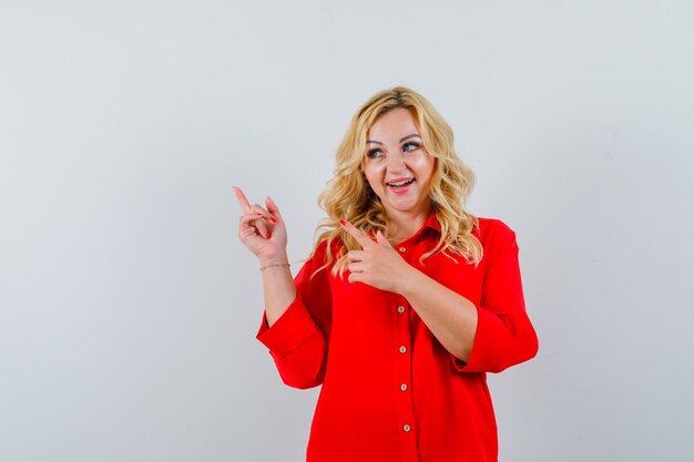 Blonde woman in red blouse pointing upper left side with index fingers and looking happy