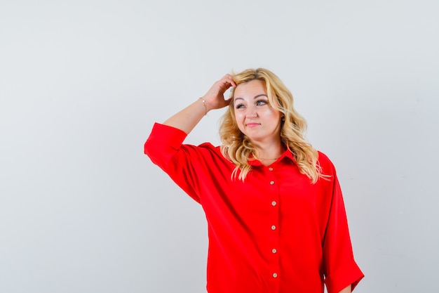 Blonde woman scratching head, thinking about something in red blouse and looking pensive