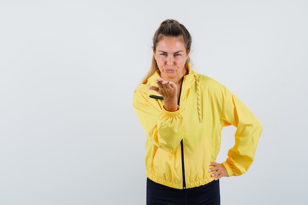 Blonde woman sending kisses to front while holding hand on waist in yellow bomber jacket and black pants and looking serious