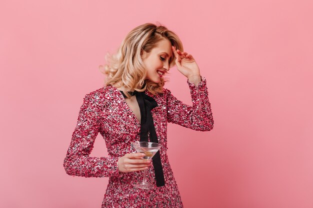 Blonde woman in shining outfit is holding martini and smiling