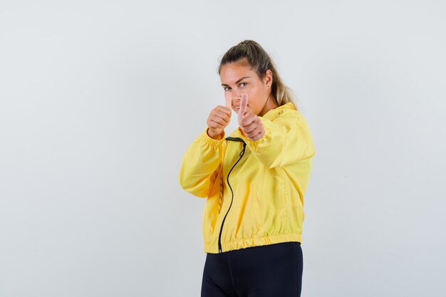 Blonde woman showing double thumbs up in yellow bomber jacket and black pants and looking happy