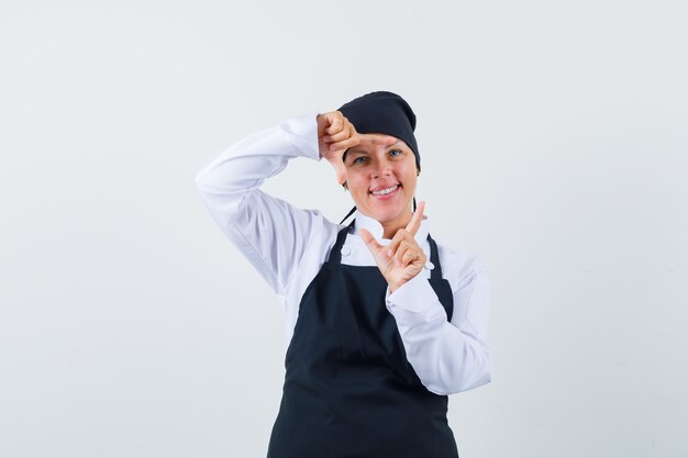 Blonde woman showing frame gesture with hands in black cook uniform and looking pretty , front view.