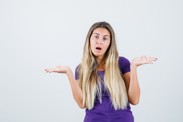 blonde woman showing helpless gesture in violet t-shirt and looking confused. front view.