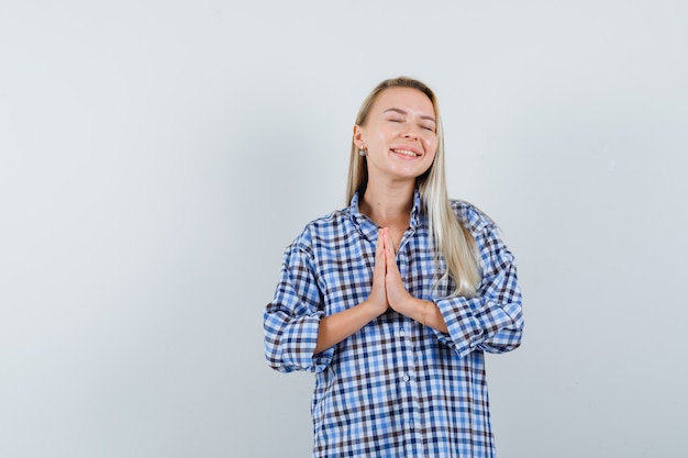 Blonde woman showing namaste gesture in blue gingham checked shirt and looking radiant. front view.