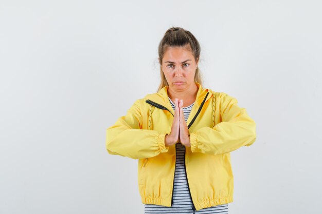 Blonde woman showing namaste gesture in yellow bomber jacket and striped shirt and looking relaxed