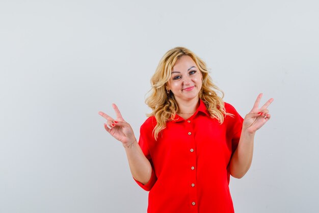 Blonde woman showing peace sign with both hands in red blouse and looking happy.