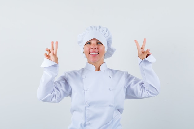 Blonde woman showing peace signs with both hands in white cook uniform and looking pretty