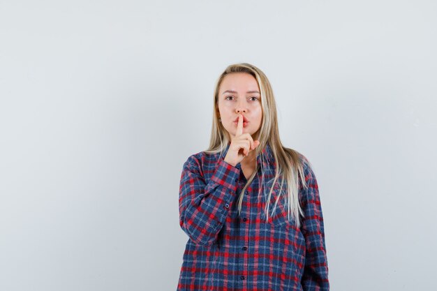 Blonde woman showing silence gesture in checked shirt and looking serious. front view.