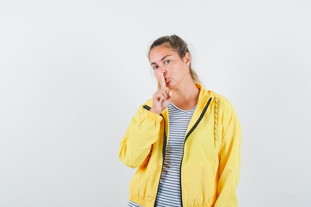 Blonde woman showing silence gesture in yellow bomber jacket and striped shirt and looking focused