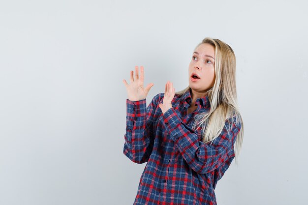 Blonde woman showing stop sign in checked shirt and looking surprised , front view.