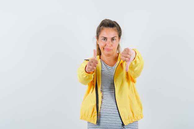 Blonde woman showing thumb up and down while grimacing in yellow bomber jacket and striped shirt and looking pensive