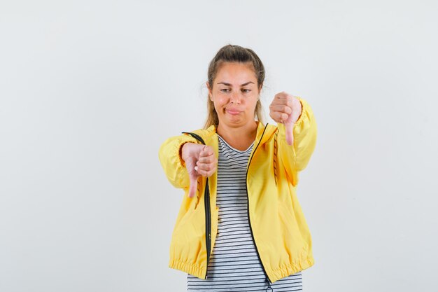 Blonde woman showing thumb up and down while grimacing in yellow bomber jacket and striped shirt and looking pensive