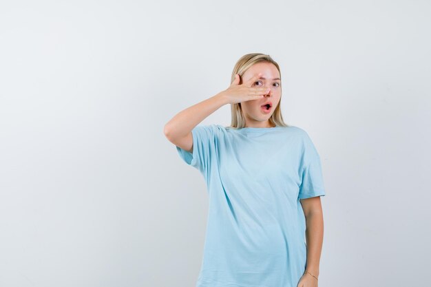 Blonde woman showing v sign on eye in blue t-shirt and looking surprised