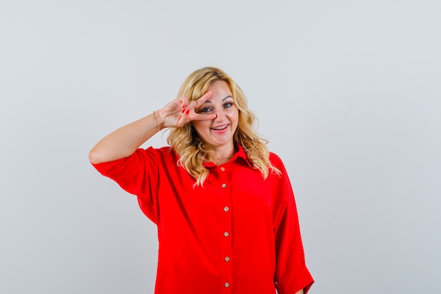 Blonde woman showing v sign on eye in red blouse and looking happy.