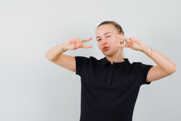 Blonde woman showing v-sign with her both hands and winking in black t-shirt and looking happy