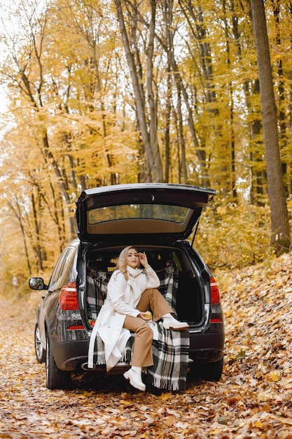 Blonde woman sitting in a black car trunk in autumn forest. Woman wearing white coat. Girl sitting on a plaid blanket.