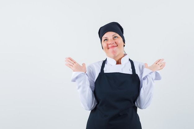 Donna bionda che diffonde le palme come mostra gesto impotente in uniforme nera del cuoco e che sembra felice, vista frontale.