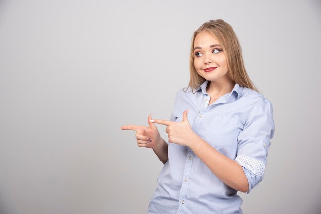 Blonde woman standing and pointing fingers aside against gray wall .