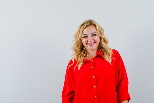 Blonde woman standing straight and posing at camera in red blouse and looking happy.