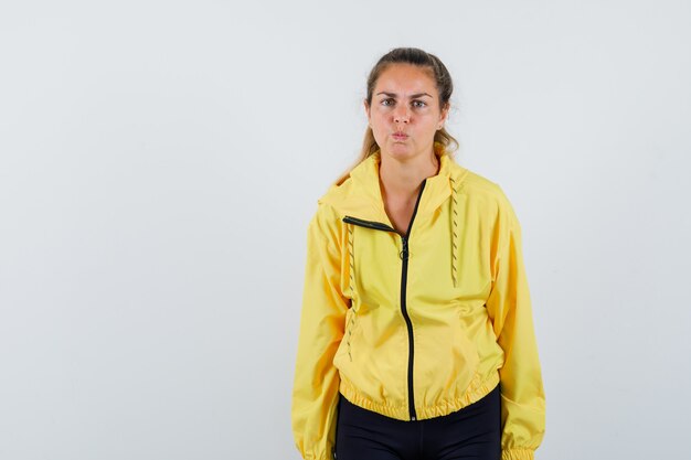 Blonde woman standing straight, puffing cheeks and posing at front in yellow bomber jacket and black pants and looking serious