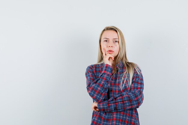 Blonde woman standing in thinking pose in checked shirt and looking serious , front view.